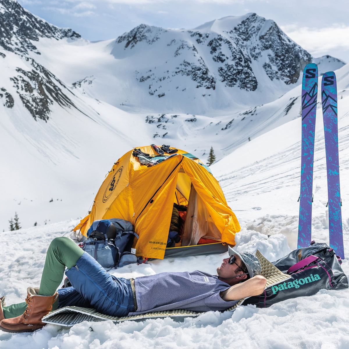 Skier resting on a Patagonia duffel beside a yellow tent at a snowy mountain camp